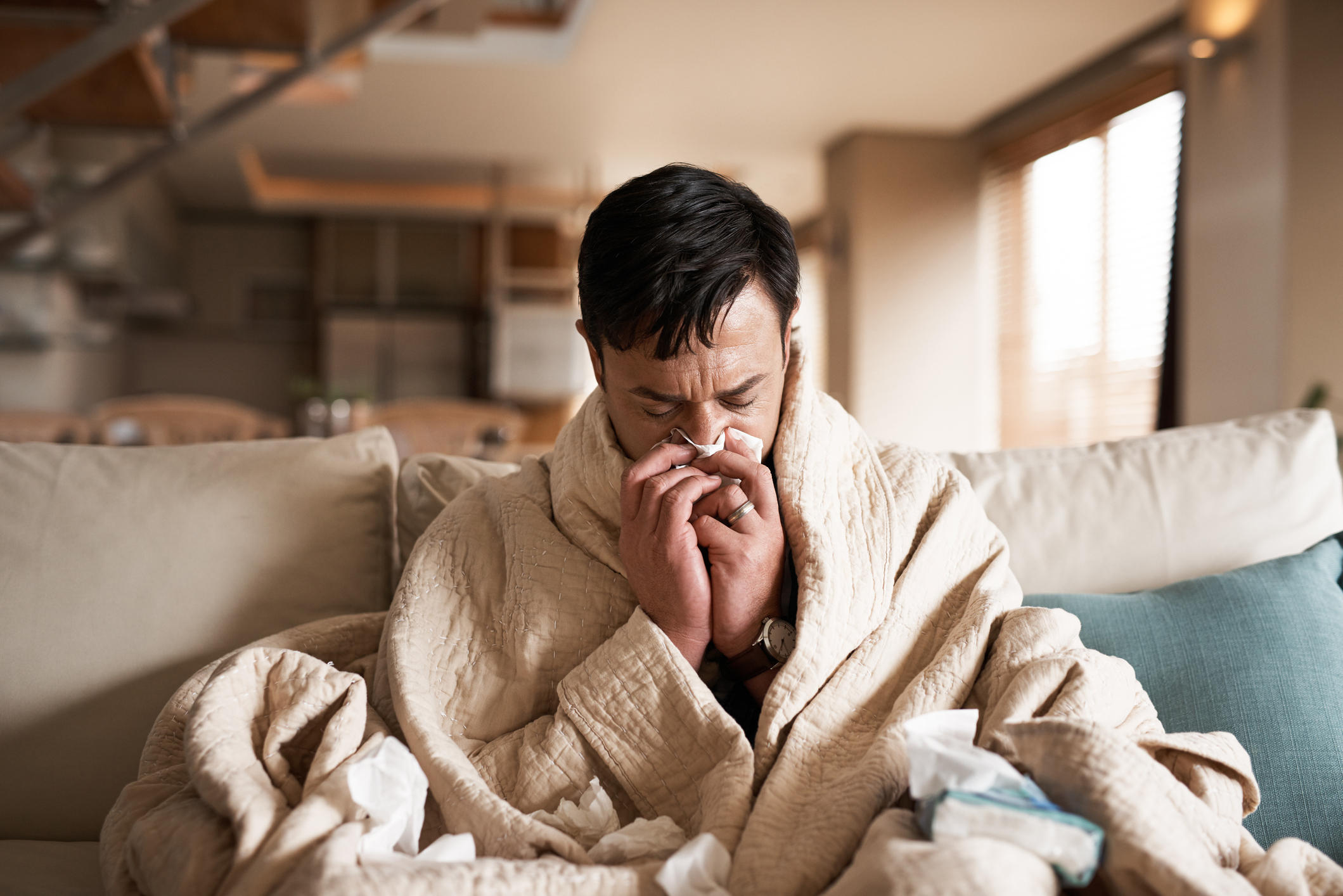 Cropped shot of a young man suffering with flu while sitting wrapped in a blanket on the sofa at home