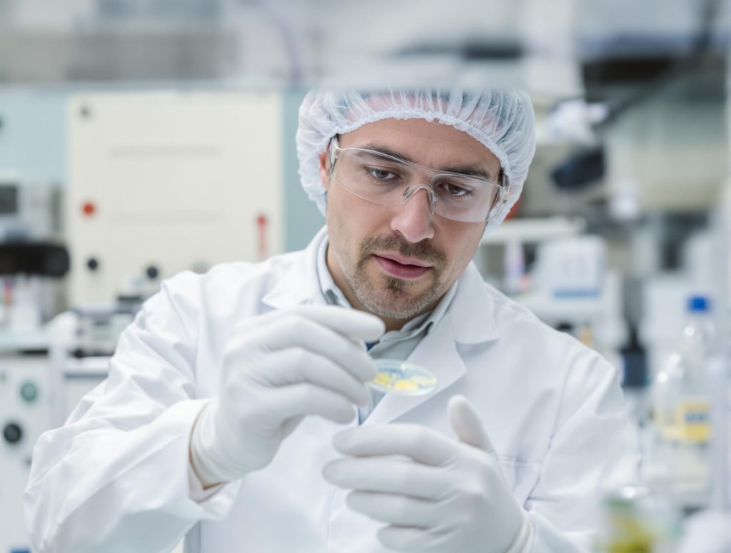 A scientist wearing a white lab coat, hairnet, safety glasses, and gloves examines a petri dish in a laboratory setting.
